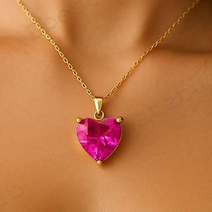 Può includere: Una collana dorata con un ciondolo a forma di cuore in pietra preziosa rosa. Il ciondolo è sfaccettato e cattura la luce, creando un effetto scintillante. La catena è delicata. Un gioiello.