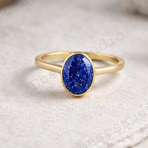 Op de afbeelding: Een gouden ring met een ovale, diepblauwe edelsteen. De ring ligt op een witte doek, met een vage achtergrond. De edelsteen heeft een gespikkelde uitstraling en de ringband is dun en eenvoudig.