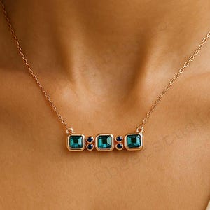 May include: A delicate gold-toned necklace featuring three square teal gemstones and two small dark blue gemstones. The necklace is displayed on a person's neck, showcasing its elegant design and color contrast.