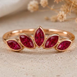 May include: A rose gold ring featuring five marquise-cut red gemstones arranged in a fan-like crown shape. The band is thin and polished, with the central stone being the largest, flanked by two smaller stones on each side in a symmetrical design.