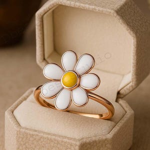 Puede incluir: Un anillo dorado con un diseño de flor de margarita. La flor tiene pétalos blancos y un centro amarillo. El anillo se exhibe dentro de un joyero beige, mostrando los delicados detalles y la artesanía del anillo.