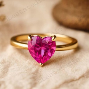 Op de afbeelding: Een gouden ring met een hartvormige, fuchsia-kleurige edelsteen. De band van de ring is van gepolijst goud en de edelsteen is gefacetteerd, waardoor het licht wordt opgevangen. De ring staat tegen een neutrale, getextureerde achtergrond.