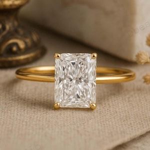 May include: A gold ring featuring a rectangular, clear gemstone. The ring has a slender band and four prongs holding the gemstone. The ring is set against a neutral-toned background.