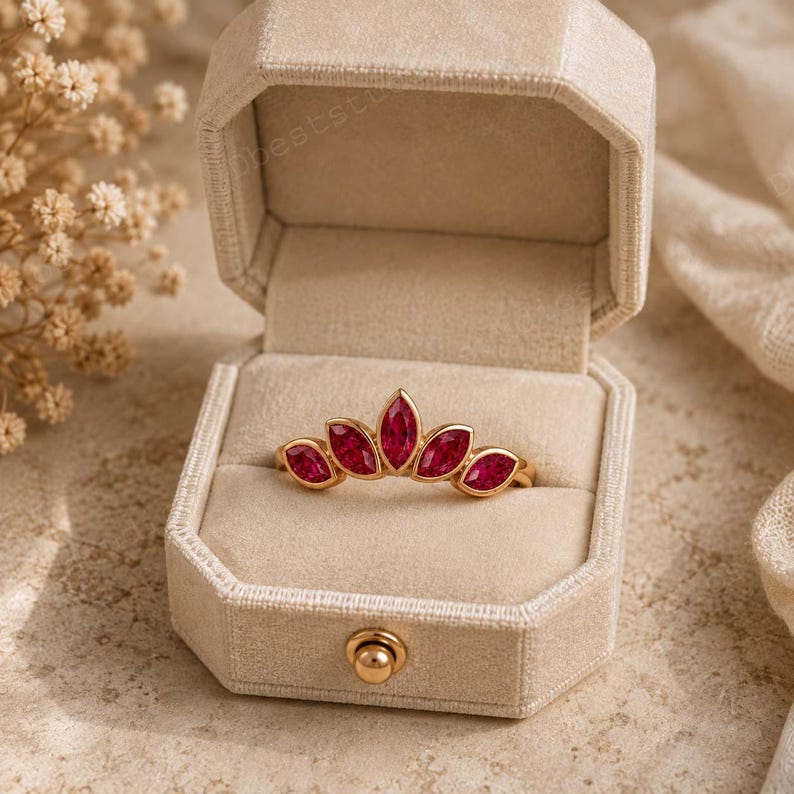 Peut inclure: Une bague en or orn&eacute;e de cinq pierres pr&eacute;cieuses rouges taille marquise dispos&eacute;es en &eacute;ventail, pr&eacute;sent&eacute;e dans un &eacute;crin hexagonal en velours beige ouvert, dot&eacute; d'un fermoir en m&eacute;tal dor&eacute; sur le bord avant.