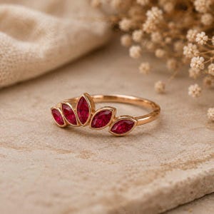 Peut inclure: Une bague en or rose orn&eacute;e de cinq rubis rouges taille marquise dispos&eacute;s en &eacute;ventail sur le dessus de l'anneau. Les pierres sont serties en ligne, le rubis central &eacute;tant le plus grand, flanqu&eacute; de deux rubis plus petits de chaque c&ocirc;t&eacute;.