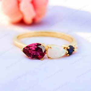 May include: A gold ring featuring a pear-shaped pink gemstone, a pear-shaped white opal, a small blue gemstone, and a row of small clear gemstones.
