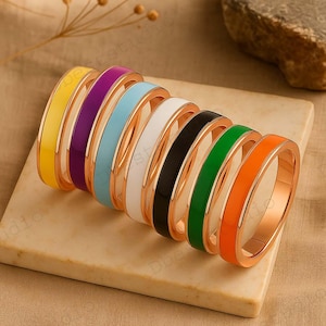 May include: Eight gold-coloured rings with enamel bands in yellow, purple, light blue, white, black, green, and orange. The rings are displayed on a light-coloured, rectangular surface.