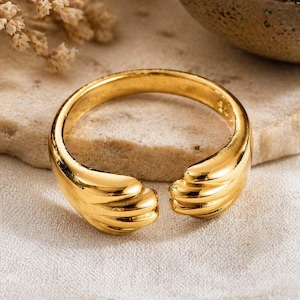 Puede incluir: Un anillo dorado con un diseño abierto. El anillo presenta un diseño texturizado y en capas en cada extremo, creando un aspecto único y elegante. El anillo probablemente está hecho de un metal brillante, que refleja la luz.