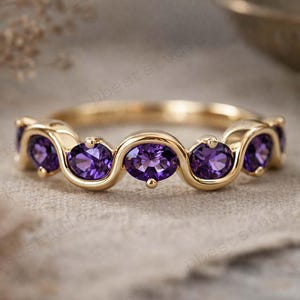 Op de afbeelding: Een gouden ring met een reeks ovaal geslepen paarse edelstenen. De edelstenen zijn gezet in een golfachtig patroon, waardoor een doorlopend ontwerp ontstaat. De ring is geplaatst tegen een neutrale achtergrond.