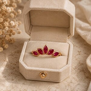 Peut inclure: Une bague en or orn&eacute;e de cinq pierres pr&eacute;cieuses rouges taille marquise dispos&eacute;es en &eacute;ventail, pr&eacute;sent&eacute;e dans un &eacute;crin hexagonal en velours beige ouvert, dot&eacute; d'un fermoir en m&eacute;tal dor&eacute; sur le bord avant.