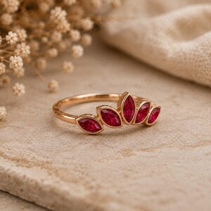 Peut inclure: Une bague en or orn&eacute;e de cinq pierres pr&eacute;cieuses rouges taille marquise dispos&eacute;es en forme d'&eacute;ventail. L'anneau est fin et poli, pr&eacute;sentant une teinte m&eacute;tallique chaude qui compl&egrave;te la couleur rouge vif et profonde des pierres facett&eacute;es.