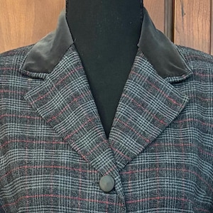 K&ouml;nnte beinhalten: Nahaufnahme eines karierten Blazers mit einem dunkelgrauen Samtkragen. Der Blazer hat ein grau-schwarz-rotes Karomuster, einen Einzelknopfverschluss und ein klassisches Revers. Das Kleidungsst&uuml;ck wird auf einer schwarzen Schaufensterpuppe pr&auml;sentiert.