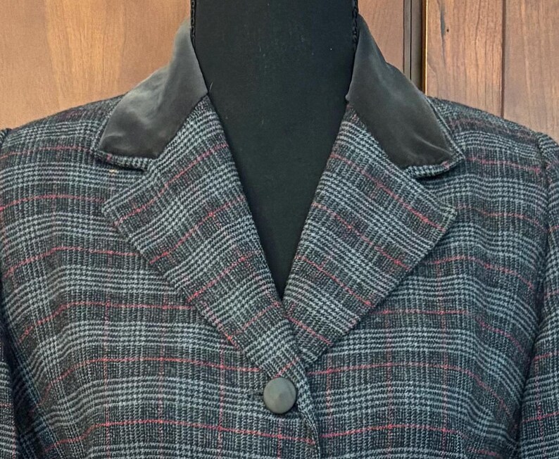 K&ouml;nnte beinhalten: Nahaufnahme eines karierten Blazers mit einem dunkelgrauen Samtkragen. Der Blazer hat ein grau-schwarz-rotes Karomuster, einen Einzelknopfverschluss und ein klassisches Revers. Das Kleidungsst&uuml;ck wird auf einer schwarzen Schaufensterpuppe pr&auml;sentiert.