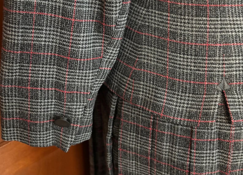 K&ouml;nnte beinhalten: Nahaufnahme eines grau-schwarz karierten Blazers mit roten Akzentlinien. Der Blazer hat eine gekn&ouml;pfte Manschette und ein ma&szlig;geschneidertes Design. Der Stoff hat ein strukturiertes Aussehen, m&ouml;glicherweise Wolle.