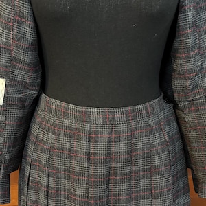 K&ouml;nnte beinhalten: Ein grauer karierter Hosenanzug mit roten Akzenten. Die Jacke hat ein Revers und wird mit einem Faltenrock kombiniert. Der Anzug hat Etiketten von "THE J.C. HILTON COMPANY" und "GLENHAVEN".