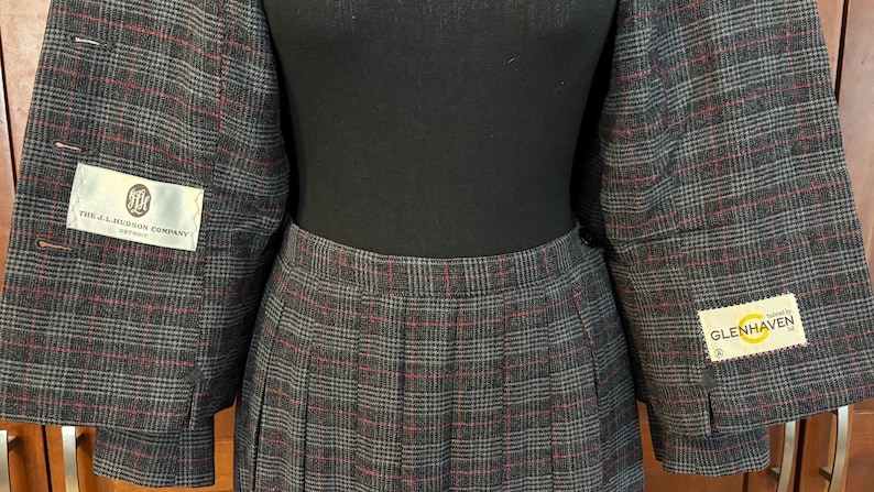 K&ouml;nnte beinhalten: Ein grauer karierter Hosenanzug mit roten Akzenten. Die Jacke hat ein Revers und wird mit einem Faltenrock kombiniert. Der Anzug hat Etiketten von "THE J.C. HILTON COMPANY" und "GLENHAVEN".