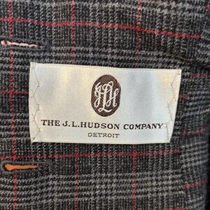 K&ouml;nnte beinhalten: Nahaufnahme einer grauen karierten Jacke mit roten Akzentlinien. Ein wei&szlig;es Etikett mit dem Text "THE J.L. HUDSON COMPANY DETROIT" und einem braunen Emblem ist in das Futter der Jacke eingen&auml;ht. Die Jacke hat einen Knopfverschluss.