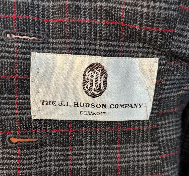 K&ouml;nnte beinhalten: Nahaufnahme einer grauen karierten Jacke mit roten Akzentlinien. Ein wei&szlig;es Etikett mit dem Text "THE J.L. HUDSON COMPANY DETROIT" und einem braunen Emblem ist in das Futter der Jacke eingen&auml;ht. Die Jacke hat einen Knopfverschluss.