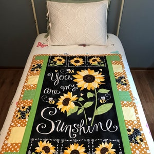 May include: A black and green quilt with a sunflower design and the text "You are my Sunshine". The quilt is made of patchwork fabric with different patterns and colors.