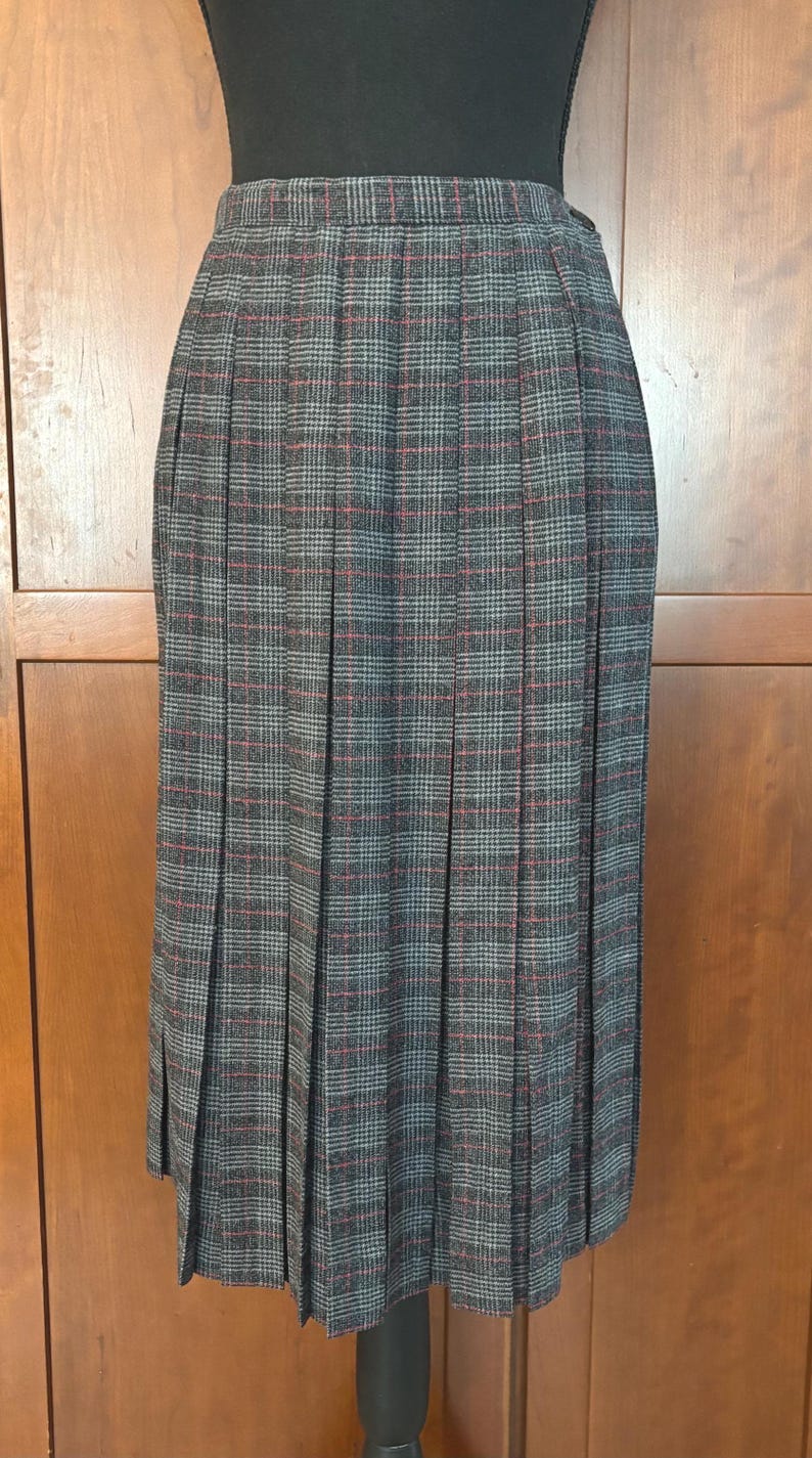 K&ouml;nnte beinhalten: Ein plissierter Rock mit grauem, roten und schwarzen Karomuster. Der Rock ist knielang und hat ein klassisches Design. Der Stoff scheint ein gewebtes Material zu sein, das f&uuml;r k&auml;lteres Wetter geeignet ist.