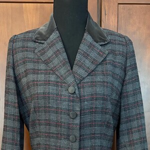 K&ouml;nnte beinhalten: Ein grau-rot-schwarz karierter Blazer mit Samtkragen. Der Blazer hat drei schwarze Kn&ouml;pfe vorne und lange &Auml;rmel. Die Jacke wird auf einer schwarzen Schaufensterpuppe pr&auml;sentiert.