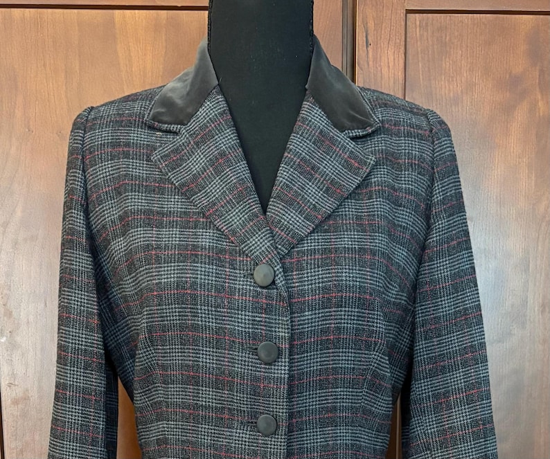 K&ouml;nnte beinhalten: Ein grau-rot-schwarz karierter Blazer mit Samtkragen. Der Blazer hat drei schwarze Kn&ouml;pfe vorne und lange &Auml;rmel. Die Jacke wird auf einer schwarzen Schaufensterpuppe pr&auml;sentiert.