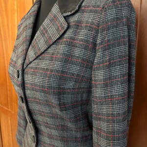 K&ouml;nnte beinhalten: Ein grauer Blazer mit rotem und wei&szlig;em Karomuster. Der Blazer hat einen dunkelbraunen Samtkragen und drei Kn&ouml;pfe. Der Stoff hat einen klassischen, ma&szlig;geschneiderten Look.