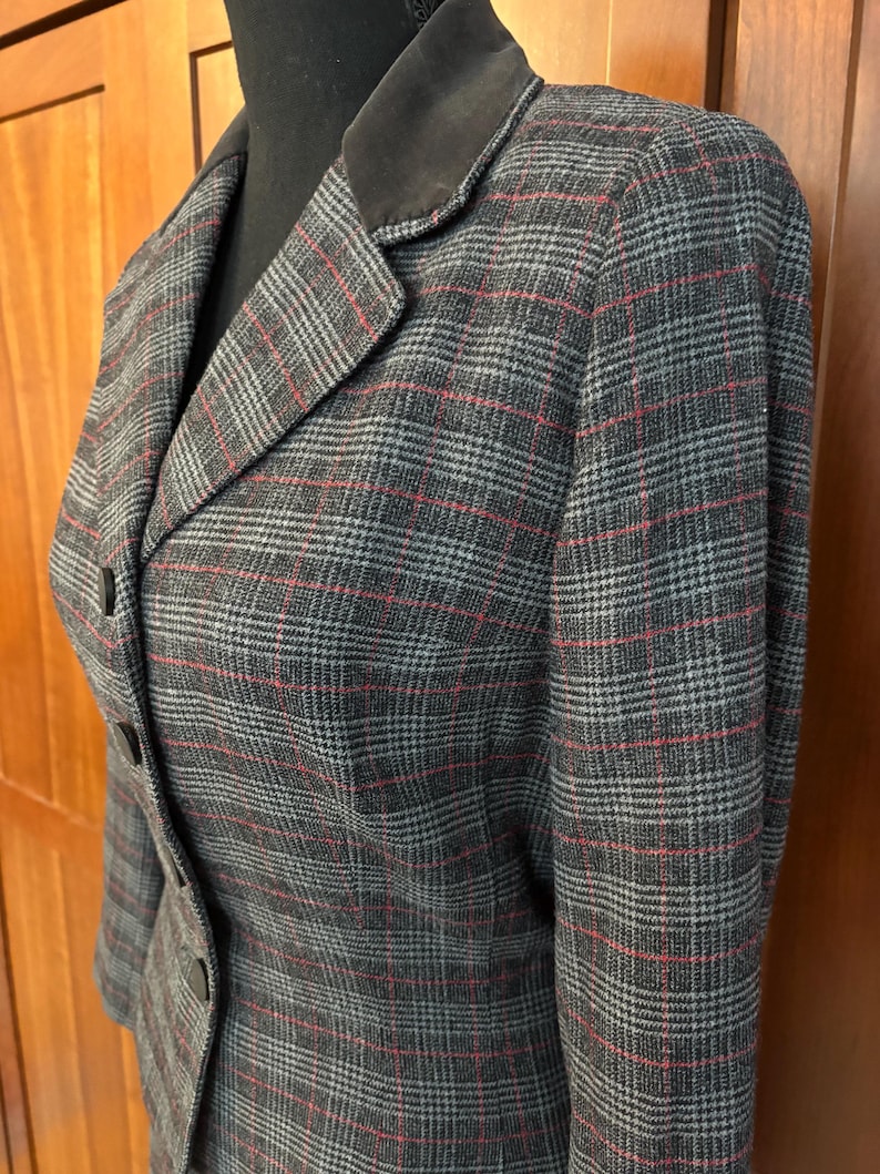K&ouml;nnte beinhalten: Ein grauer Blazer mit rotem und wei&szlig;em Karomuster. Der Blazer hat einen dunkelbraunen Samtkragen und drei Kn&ouml;pfe. Der Stoff hat einen klassischen, ma&szlig;geschneiderten Look.