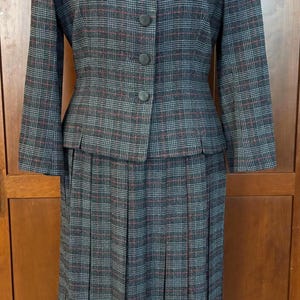 K&ouml;nnte beinhalten: Ein Vintage-Rockanzug mit grauem Karomuster. Die Jacke hat einen Reverskragen, drei Kn&ouml;pfe und lange &Auml;rmel. Der Rock ist plissiert. Der Anzug ist grau mit roten und blauen Akzenten.