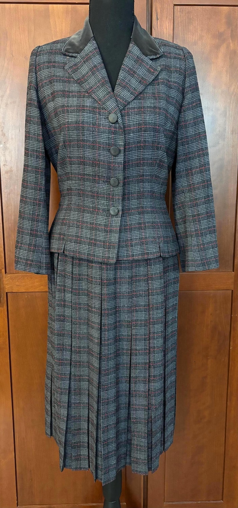 K&ouml;nnte beinhalten: Ein Vintage-Rockanzug mit grauem Karomuster. Die Jacke hat einen Reverskragen, drei Kn&ouml;pfe und lange &Auml;rmel. Der Rock ist plissiert. Der Anzug ist grau mit roten und blauen Akzenten.