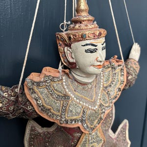 May include: A detailed shot of a traditional Asian puppet with a painted face, adorned with a gold headdress and elaborate, sequined costume in shades of red, gold, and beige. The puppet is suspended by strings.