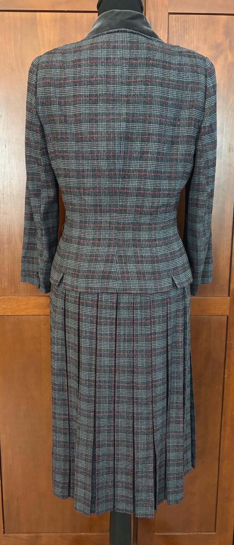 K&ouml;nnte beinhalten: Ein grauer karierter Hosenanzug mit einem Faltenrock. Die Jacke hat einen Samtkragen und der Anzug hat ein rot-blaues Karomuster. Der Anzug wird auf einer Schaufensterpuppe pr&auml;sentiert.
