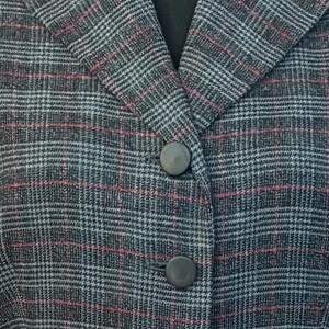 K&ouml;nnte beinhalten: Nahaufnahme eines grauen Blazer mit Karomuster und roten und wei&szlig;en Akzenten. Der Blazer hat ein fallendes Revers und zwei graue Kn&ouml;pfe. Der Stoff hat eine strukturierte Optik, m&ouml;glicherweise Wolle oder Tweed. Der Stil ist klassisch und professionell.