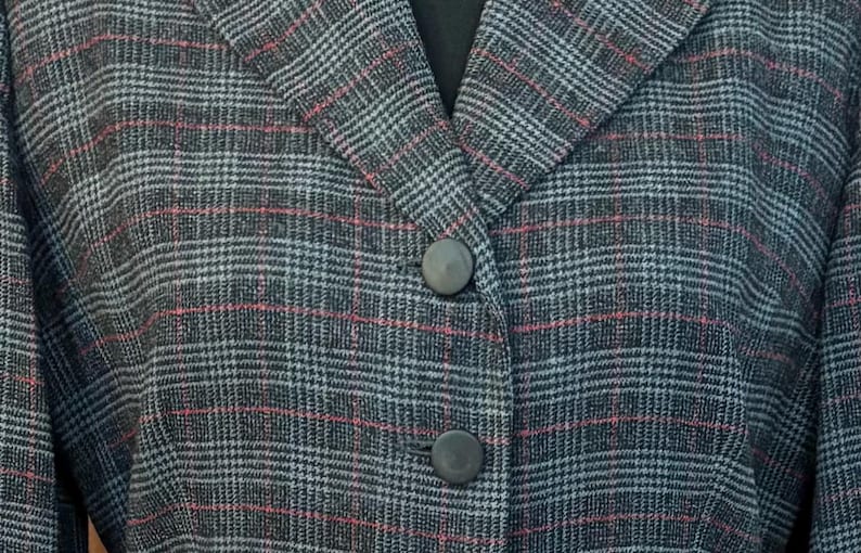 K&ouml;nnte beinhalten: Nahaufnahme eines grauen Blazer mit Karomuster und roten und wei&szlig;en Akzenten. Der Blazer hat ein fallendes Revers und zwei graue Kn&ouml;pfe. Der Stoff hat eine strukturierte Optik, m&ouml;glicherweise Wolle oder Tweed. Der Stil ist klassisch und professionell.
