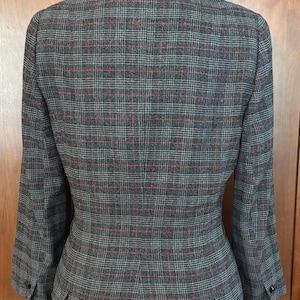 K&ouml;nnte beinhalten: Ein grauer Anzug mit Karomuster, bestehend aus einem Blazer mit Samtkragen und einem passenden Rock. Der Anzug hat ein klassisches Muster mit roten und grauen Linien. Der Blazer hat Kn&ouml;pfe an den &Auml;rmeln und eine taillierte Passform. Der Rock hat Falten.