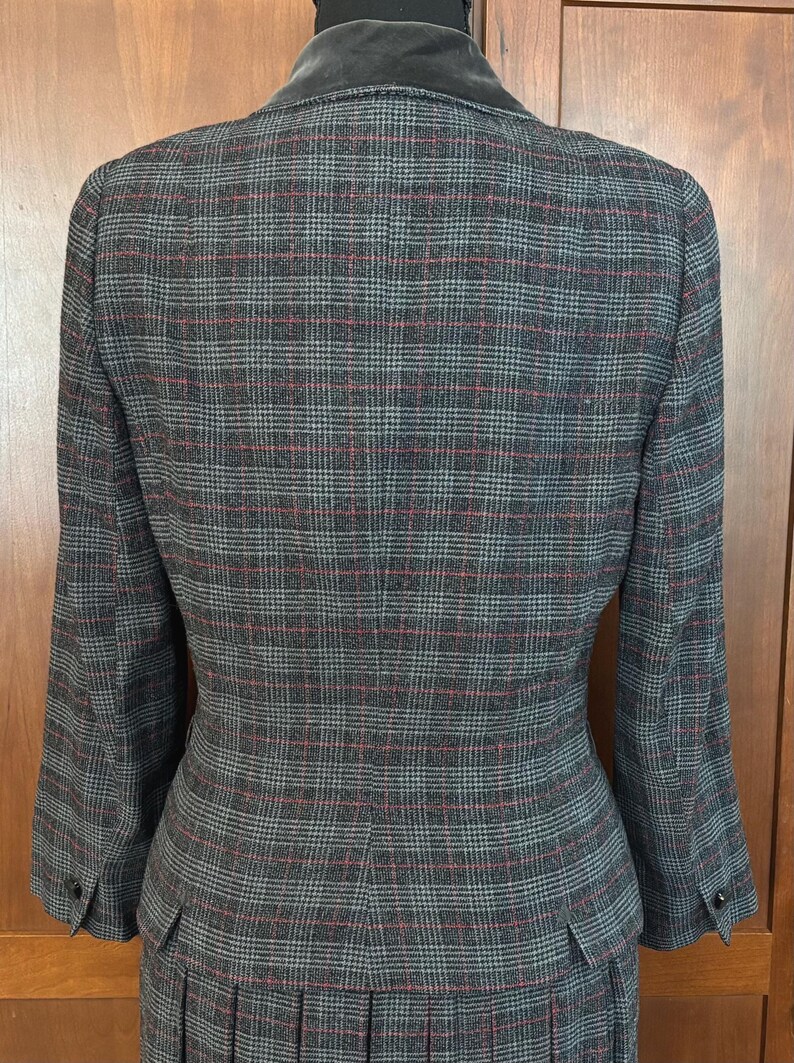 K&ouml;nnte beinhalten: Ein grauer Anzug mit Karomuster, bestehend aus einem Blazer mit Samtkragen und einem passenden Rock. Der Anzug hat ein klassisches Muster mit roten und grauen Linien. Der Blazer hat Kn&ouml;pfe an den &Auml;rmeln und eine taillierte Passform. Der Rock hat Falten.