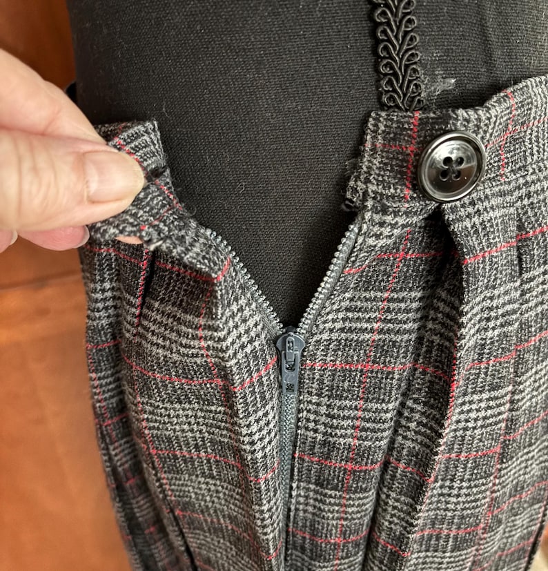 K&ouml;nnte beinhalten: Nahaufnahme eines grau karierten Rocks mit roten Akzentlinien. Der Rock hat einen Rei&szlig;verschluss und einen Knopf. Der Stoff ist &uuml;ber einer schwarzen Mannequin-Form drapiert.