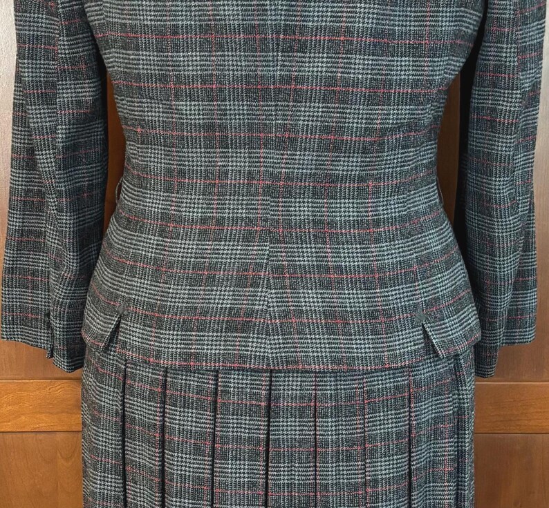 K&ouml;nnte beinhalten: Ein grauer karierter Hosenanzug mit Blazer und Faltenrock. Der Anzug hat ein klassisches Karomuster in Grau, Rot und Hellblau. Der Blazer hat eine taillierte Passform mit Taschen, und der Rock ist plissiert.