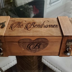 May include: A wooden box with a natural finish, featuring the engraved text "The Bendrosses" on the lid. A heart shape with the letter "B" is carved on the front. The box has metal latches and hinges.