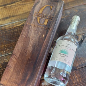 Può includere: Una bottiglia di Tequila Casamigos Blanco accanto a una scatola di legno. La scatola è incisa con il nome "GRAHAM" e "ESTABLISHED 2022". La bottiglia di tequila ha un'etichetta bianca con testo e un logo turchese.