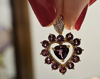 Puede incluir: Pendiente dorado con un colgante en forma de corazón. El colgante presenta una piedra preciosa morada en forma de corazón en el centro, rodeada de piedras moradas más pequeñas. La parte superior del pendiente tiene un pequeño detalle con diamantes.