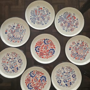 Peut inclure: Un ensemble de sept assiettes rondes en émail blanc cassé ornées d'illustrations folkloriques complexes en rouge et bleu. Les motifs incluent des coqs stylisés, des voiliers, des fleurs et des personnages en tenue traditionnelle, avec des bords sombres.