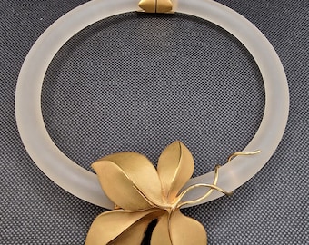 May include: A frosted glass collar necklace with a gold-tone floral pendant. The necklace has a hinged clasp and a large, stylized flower with multiple petals. The design is elegant and modern, suitable for special occasions.