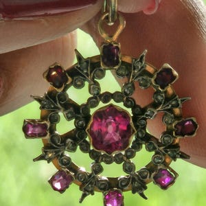 Op de afbeelding: Een vintage hanger met een goudkleurige zetting, met een centrale, grote, roze edelsteen omringd door kleinere roze edelstenen en sierlijke details. De hanger wordt omhoog gehouden door een goudkleurige lus.