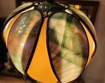 May include: A decorative table lamp with a stained glass shade. The shade features a floral design with shades of green, purple, and yellow. The base and stem are made of bronze-coloured metal. The lamp is lit, casting a warm glow.