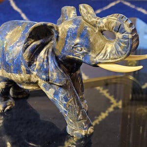 Op de afbeelding: Een blauw en gouden gegraveerd olifantenbeeldje met ivoren slagtanden. De olifant staat op een zwart oppervlak.