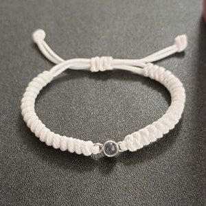 May include: A white braided bracelet with a small, round, dark blue gemstone in the center.