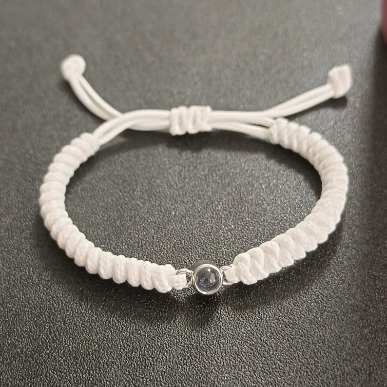 May include: A white braided bracelet with a small, round, dark blue gemstone in the center.