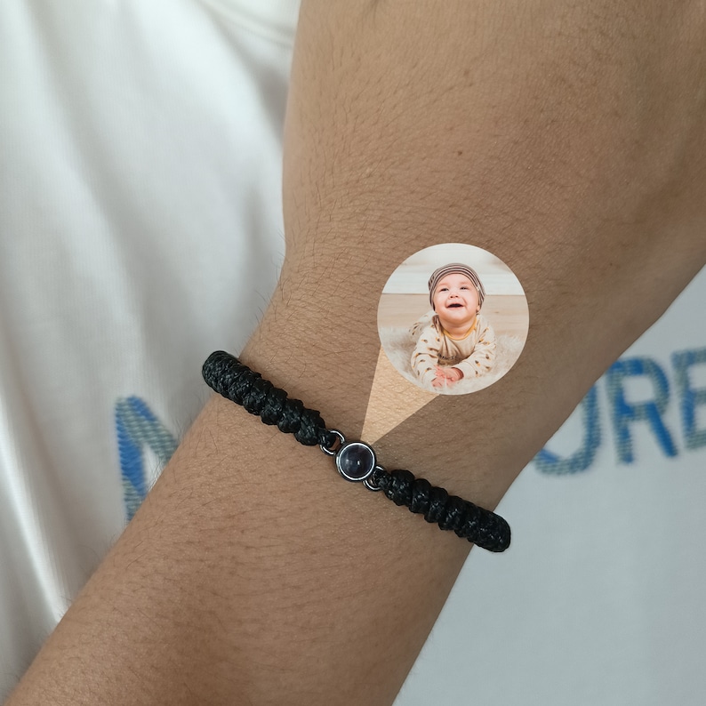 May include: A black braided bracelet with a small round black charm featuring a photo of a baby. The bracelet is worn on a person's wrist.