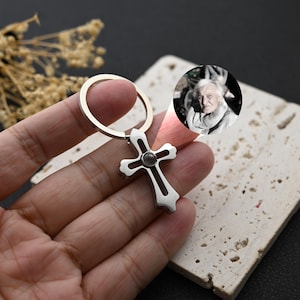 Puede incluir: Llavero plateado en forma de cruz con un llavero circular. La cruz tiene un centro oscuro y una pequeña incrustación redonda. Una pequeña imagen circular de una persona es visible en el fondo.